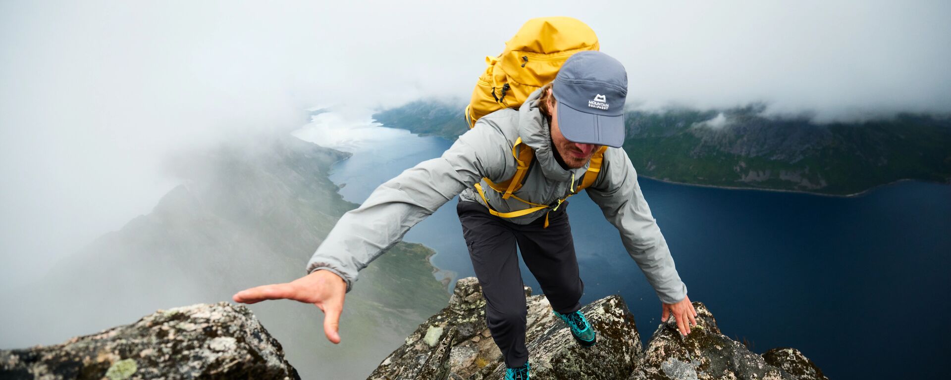 Ein Bergsteiger in einer grauen Aerotherm Jacke von Mountain Equipment hoch oben über einem Fjord in Norwegen.