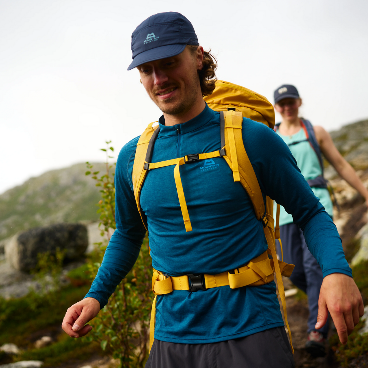 Ein Wanderer, der das dunkelblaue Longsleeve Ignis von Mountain Equipment mit einem gelben Rucksack trägt und sich schnell nach vorne bewegt.