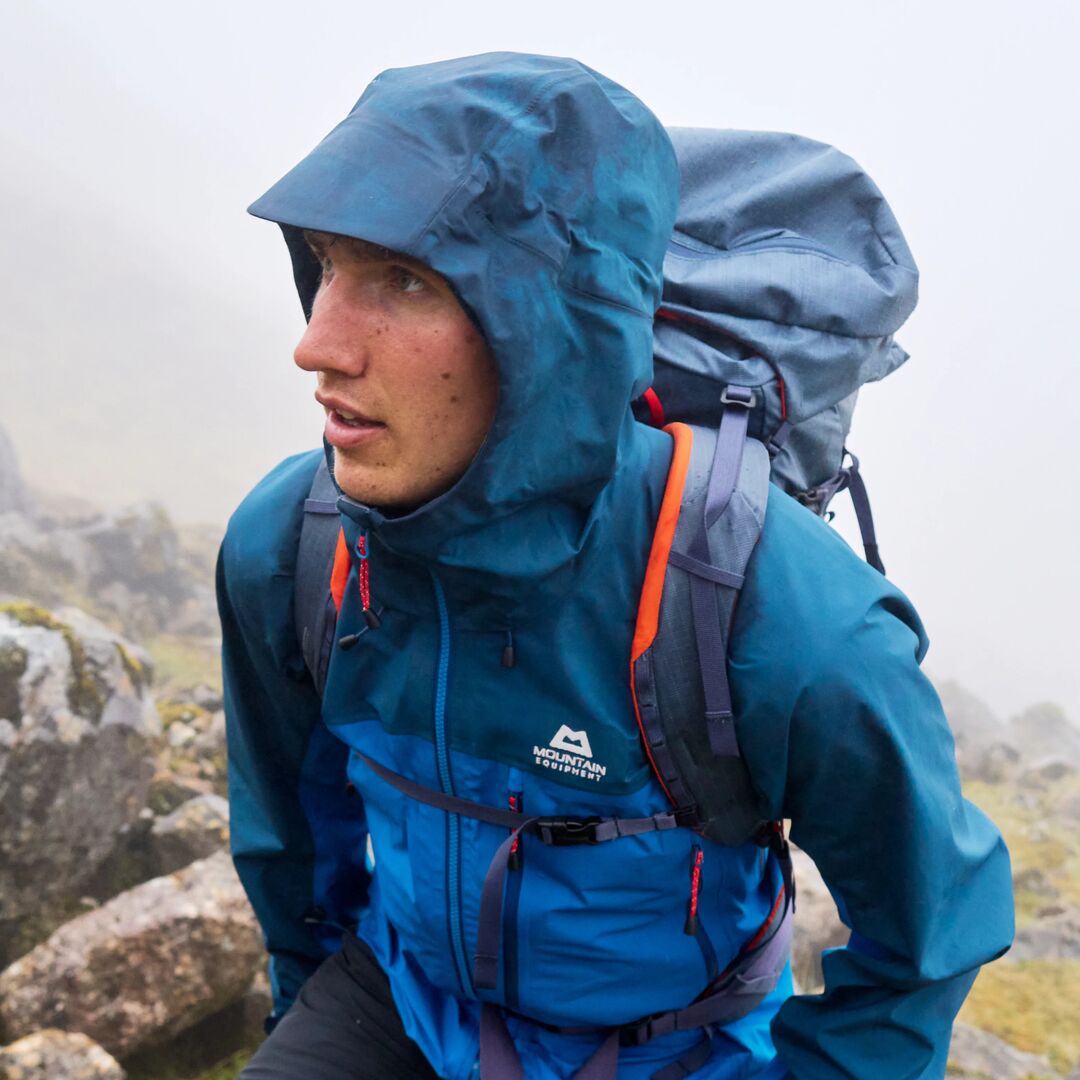 Ein Wanderer, der eine blaue Hardshelljacke von Mountain Equipment bei Regen und Nebel am Berg trägt. 
