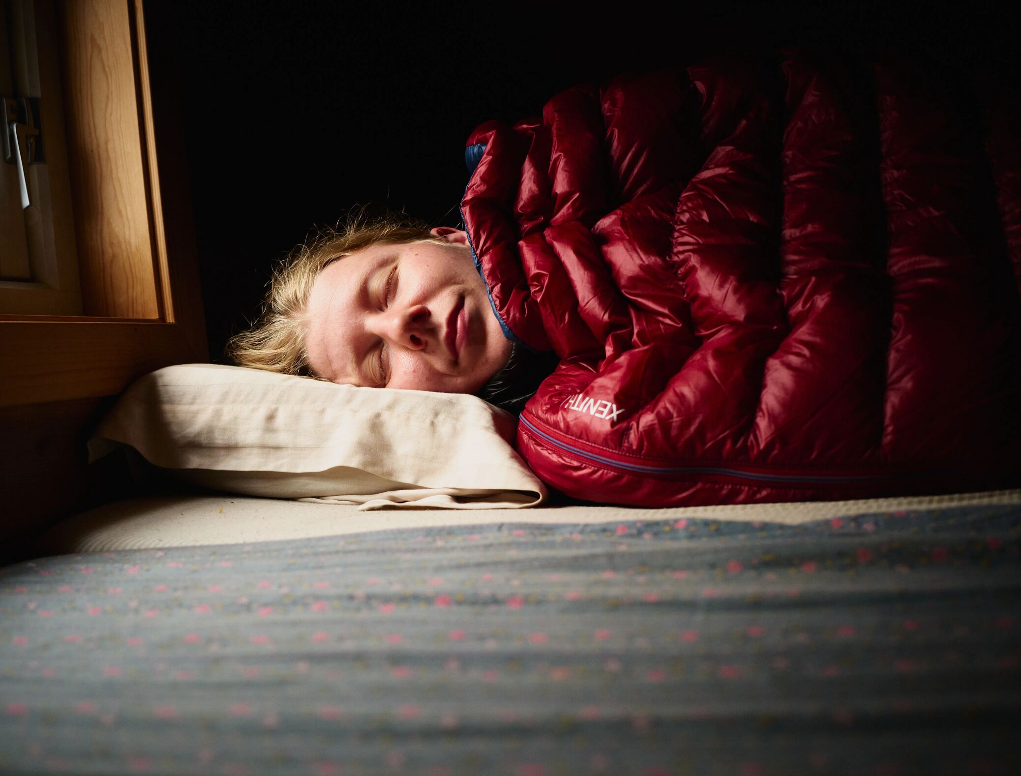 Eine junge blonde Frau, die in einem roten Xenith Schlafsack von Mountain Equipment in einer Berghütte schläft.