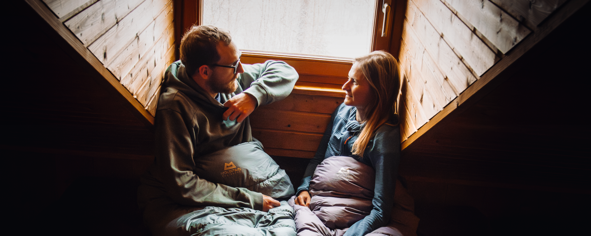 Ein junger Mann und eine junge Frau die in Classic 350 Daunenschlafsäcken von Mountain Equipment in einer Berghütte sitzen.