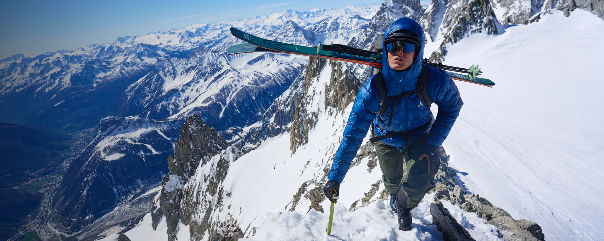 Ein Bergsteiger auf Skitour mit einer blauen Xenith Hooded Jacket von Mountain Equipment auf einem verschneiten Gipfel.