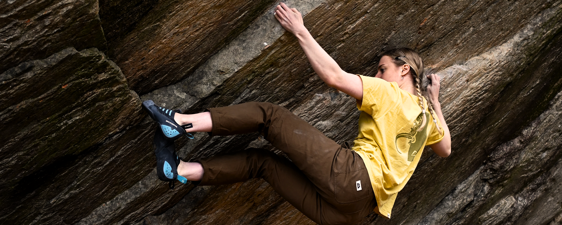 Eine junge Frau beim Bouldern an einem Fels. Dabei trägt sie das gelbe Podus Tee sowie die braune Flowstone Pant von Mountain Equipment aus der Stonewear Kollektion.